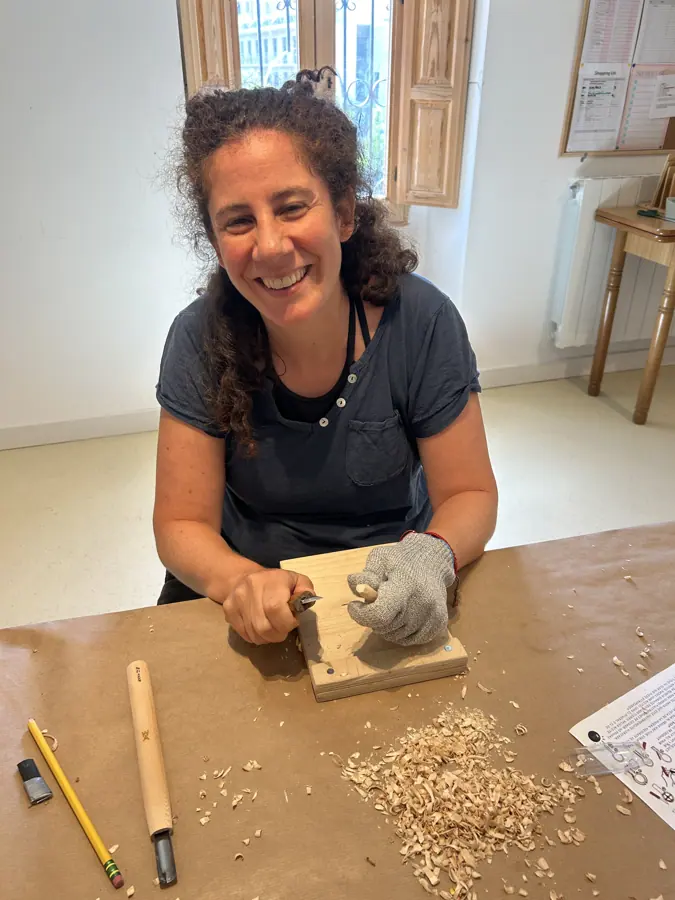 a participant smiling as she carves a spoon blank, shavings on the bench